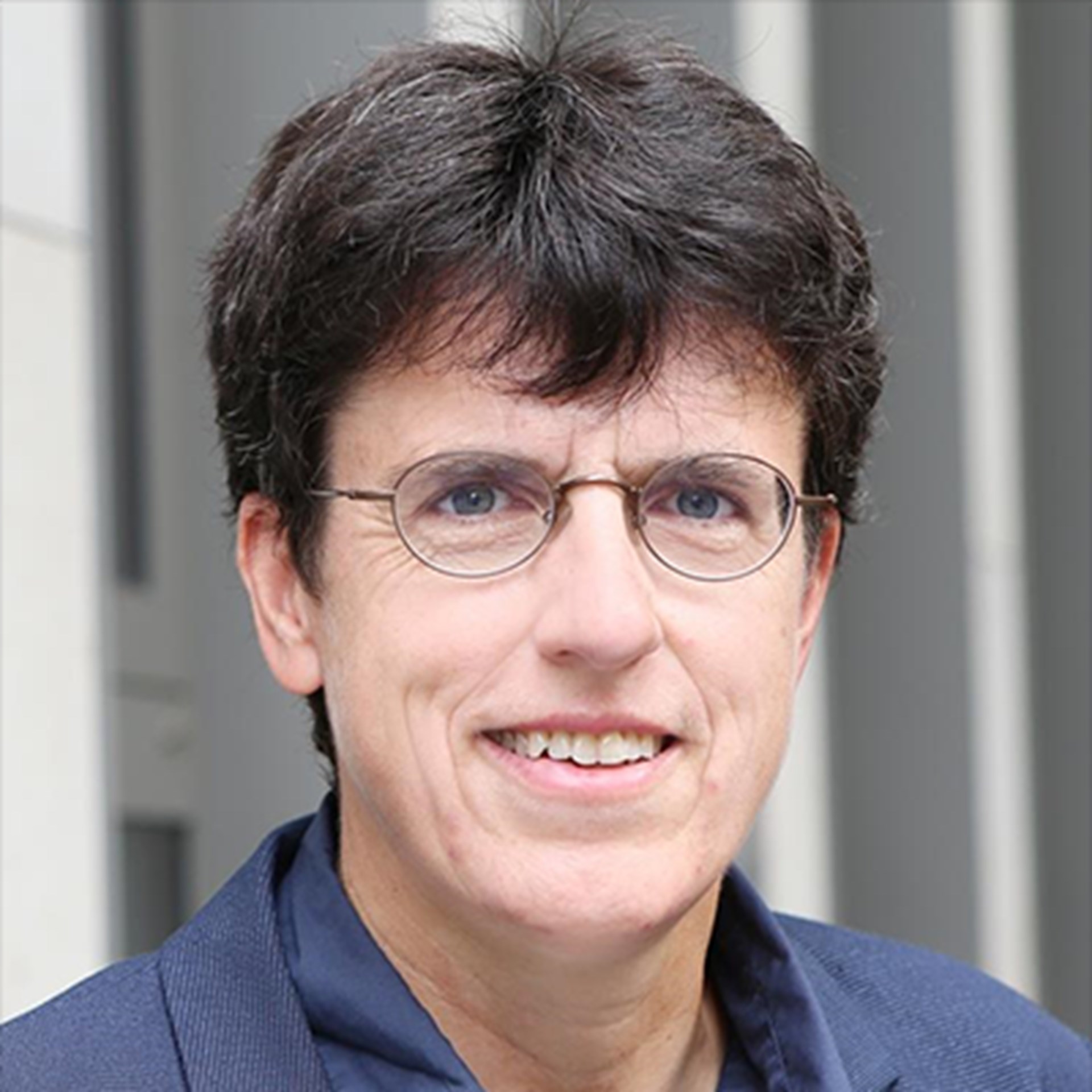 Lächelnde Frau mit schwarzen Haaren und Brille, die ein blaues Hemd mit einer blauen Anzugjacke trägt und vor einem hellen Gebäude steht