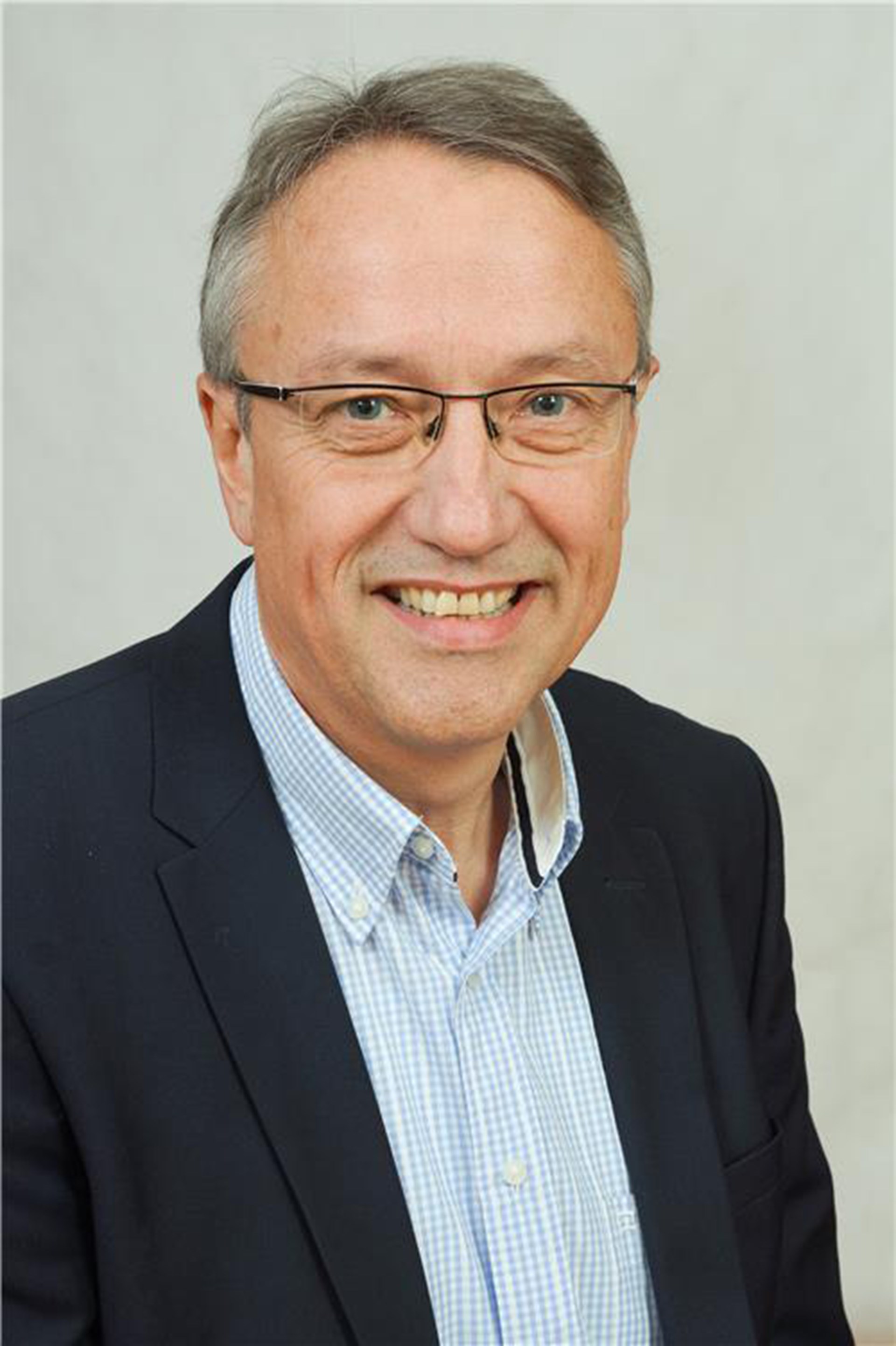 Lächelnder Mann mit grauen Haaren und Brille, der ein blaues Hemd und eine dunkelblaue Anzugjacke trägt